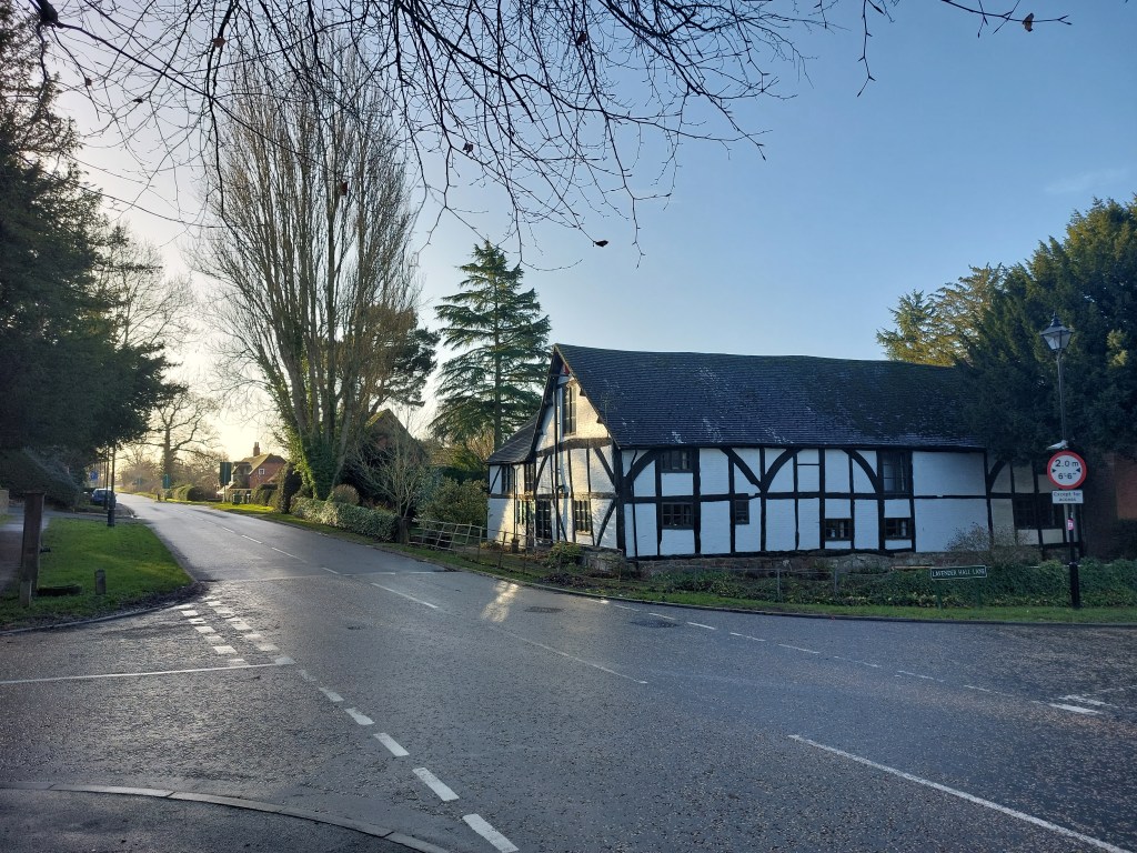 Photo shows a black and white timbered, two-storey house to the right of a tarmacked crossroads against a clear blue sky