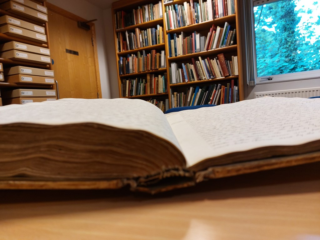 Closeup of the bottom of a large manorial court book, oblique so you can't read the text, with bookshelves in the background
